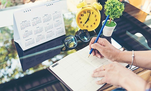 Take time a person writing on a calendar with a clock and flowers on a desk
